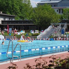 Schwimmerbecken mit rot-weißen-Fähnchen dekoriert. Im Hintergrund sieht man die Freibad-Rutsche und Personen, die im Wasser sind oder auf der Liegewiese entspannen.