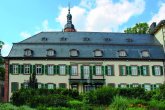 Das Haus Rose liegt direkt am Rheinufer. Ein Fachwerkhaus mit grünen Fensterläden und einen Balkon in der Mitte. Davor wächst eine Hecke und dahinter sieht man die Spitze des Kirchturms.