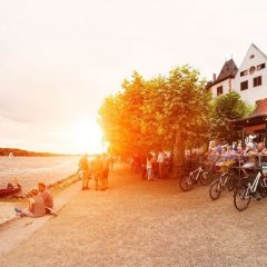 Personen sitzen am Weinstand, andere sitzen direkt am Rheinufer oder gehen dort spazieren. Im Hintergrund geht die Sonne hinter den Platanenbäumen unter.