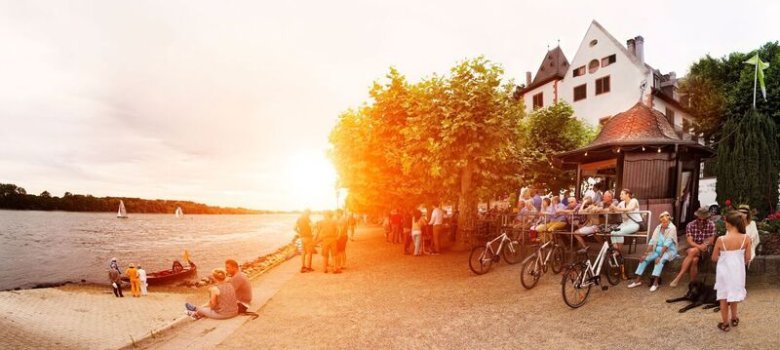 Personen sitzen am Weinstand, andere sitzen direkt am Rheinufer oder gehen dort spazieren. Im Hintergrund geht die Sonne hinter den Platanenbäumen unter.