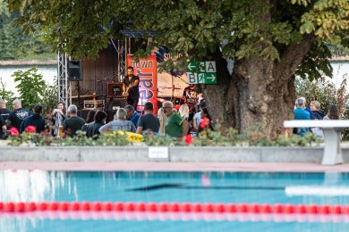 Im Vordergrund sieht man das Becken des Freibads, dahinter sieht man Zuschauer, die den Aufttritt einer Band auf einer Bühne verfolgen.