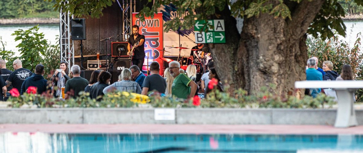 Der Sound of Eltville findet Open-Air im Rosenbad statt. Im Vordergrund sieht man das Becken des Freibads, dahinter sieht man Zuschauer, die den Aufttritt einer Band auf einer Bühne verfolgen.