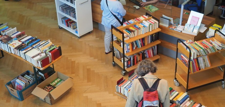 Bückerflohmarkt in der Mediathek: Eine Frau stöbert in den Büchern, eine andere Dame unterhält sich mit einer Mediatheksmitarbeiterin an der Ausleihe.