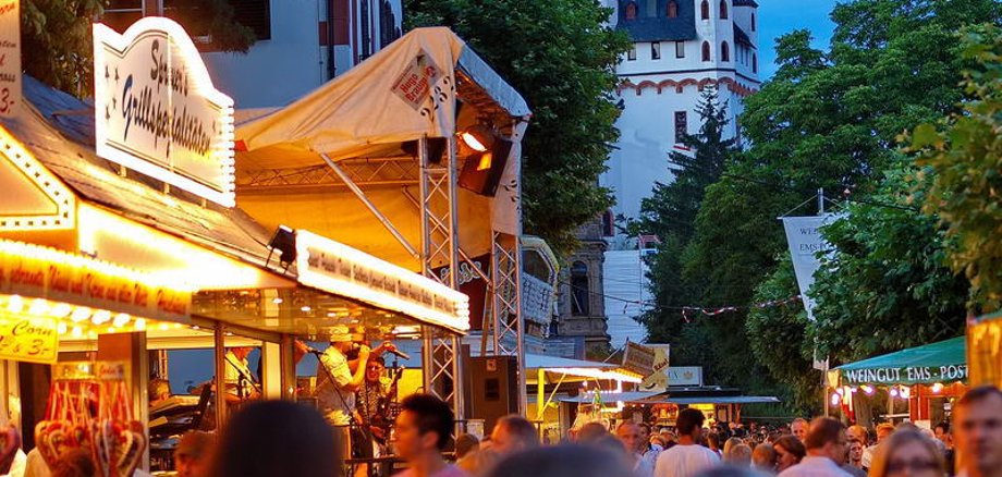 Menschenmassen schlendern entlang von aufgebauten Buden, im Hintergrund leuchtet die Kurfürstliche Burg.