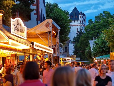 Menschenmassen schlendern entlang von aufgebauten Buden, im Hintergrund leuchtet die Kurfürstliche Burg.