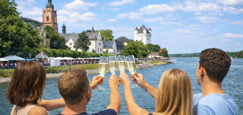 Vier Menschen von hinten zu sehen, stoßen mit Sekt an und blicken über den Rhein in Richtung Eltviller Uferpromenade