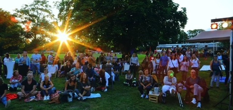 Publikum sitzt auf der Wiese, im Hintergrund scheint die Sonne durch die Bäume.