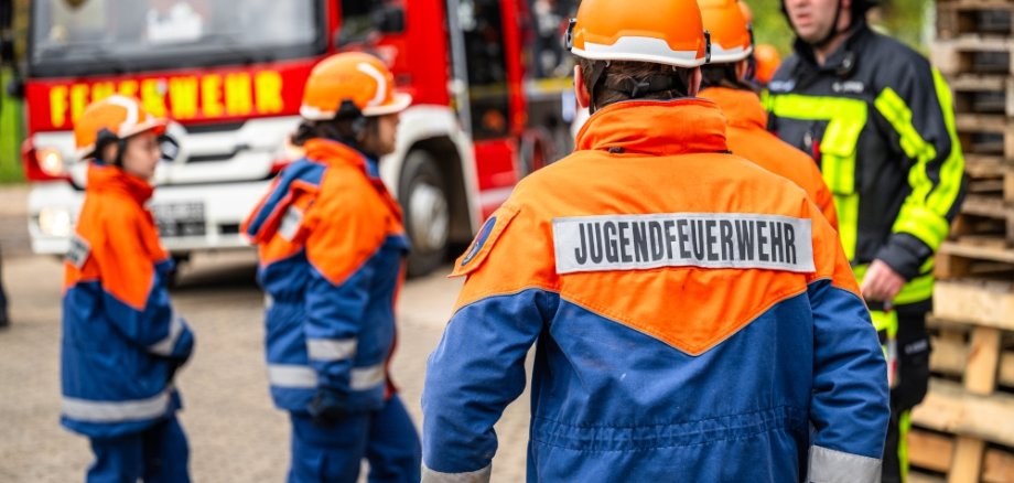 Mehrere Jungendliche in Ausrüstung von hinten zu sehen, im Hintergrund steht ein Feuerwehrauto