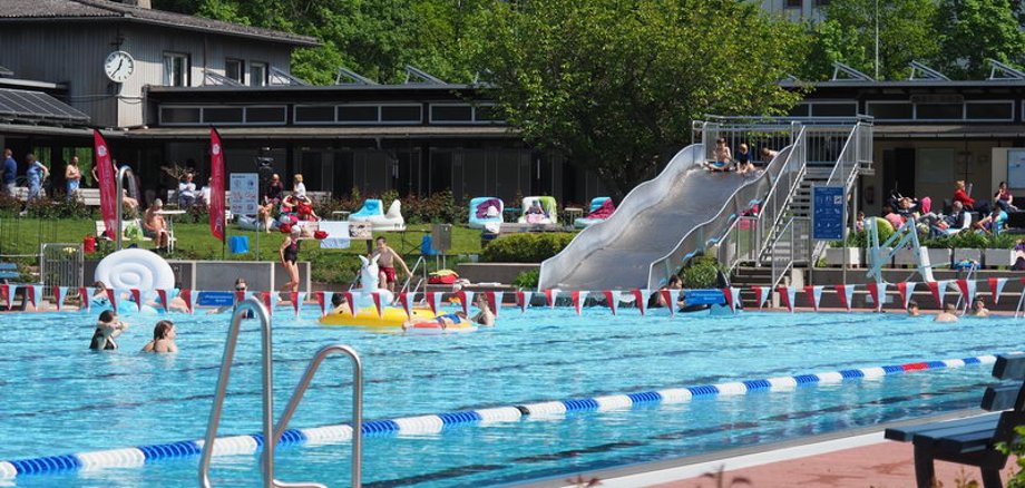 Schwimmerbecken mit rot-weißen-Fähnchen dekoriert. Im Hintergrund sieht man die Freibad-Rutsche und Personen, die im Wasser sind oder auf der Liegewiese entspannen.