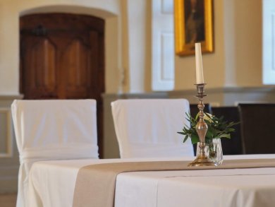 Jede Hochzeit ist individuell. Zwei Stühle stehen an einem Tisch, auf dem sich eine weiße Tischdecke, ein kleines Rosengesteck und eine Kerze befinden.
