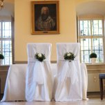 Zwei Stühle sind weiß bezogen und mit Blumen geschmückt, im Hintergrund hängt ein Gemälde.