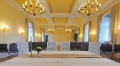 Der Kurfürsten Saal bietet ein festliches Ambiente. Foto aus Sicht der Standesbeamtin in den ERaum: Vorne stehen zwei Stühle für das Brautpaar, dahinter stehen Stuhlreihen und goldene Kronleuchter hängen von der Decke.