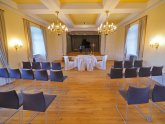 Blick in den Kurfürstensaal. Im Kurfürstensaal stehen mehrere Stuhlreihen, Kronleuchter hängen von der Decke und vorne steht ein weiß überzogener Tisch mit Stühlen für das Brautpaar.