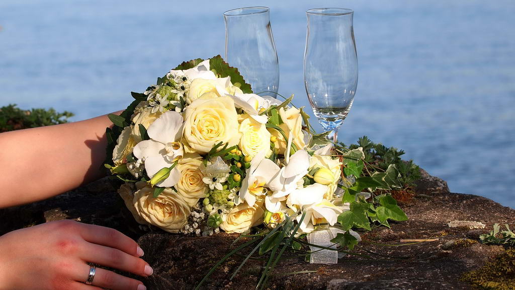 Feiern Sie den Bund der Ehe mit Blick auf den Rhein. Eine Damenhand mit Ring hält einen weißen Blumenstrauß, davor stehen zwei Gläser Sekt auf einer Mauer, im Hintergrund liegt der Rhein.