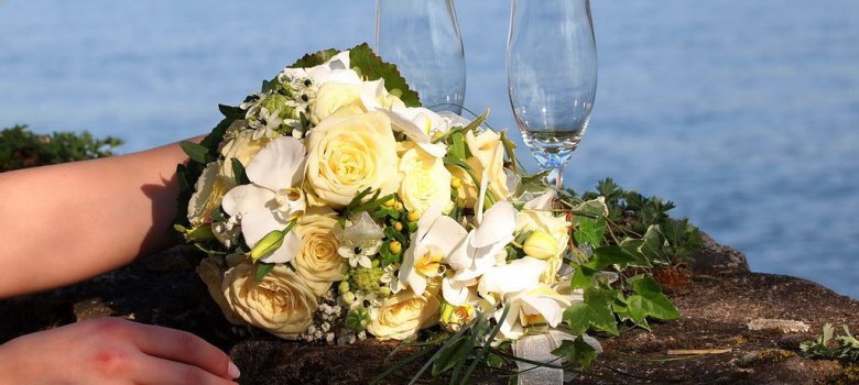 Feiern Sie den Bund der Ehe mit Blick auf den Rhein. Eine Damenhand mit Ring hält einen weißen Blumenstrauß, davor stehen zwei Gläser Sekt auf einer Mauer, im Hintergrund liegt der Rhein.
