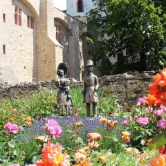 Das bronzene Biedermeierpaar steht im blühenden Rosenbeet vor der Kurfürstlichen Burg.