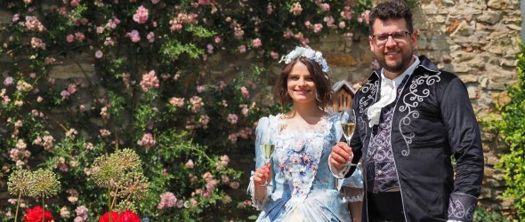 Das Eltviller Rosenpaar, Julia und Valentin Schmitt, ist festlich im Biedermeierstil gekleidet und steht mit einem Glas Sekt vor Rosen in der Kurfürstlichen Burg.