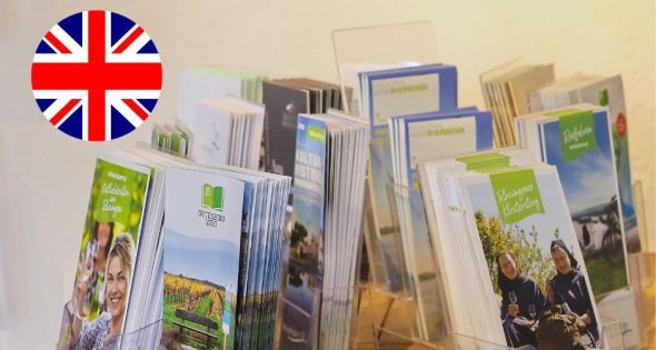 Mehrere Flyer stehen auf einem Tisch in durchsichtigen Ständern, oben links in der Ecke ist ein englischen Flagge als Ikon eingefügt.