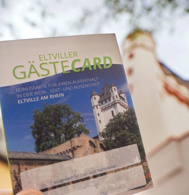 Die Eltviller GästeCard bietet Vergünstigungen für Übernachtungsgäste. Die Eltviller GästeCard, im Hintergrund sieht man den Turm der Kurfürstlichen Burg Eltville.
