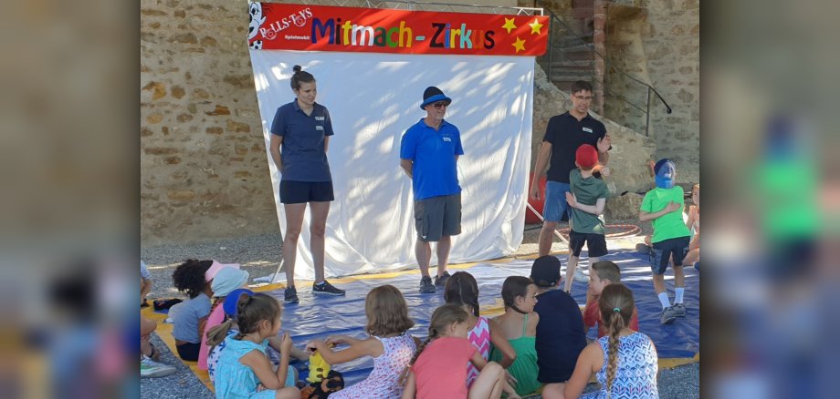 Kinder sitzen um eine kleine Bühne herum, auf der drei Erwachsene stehen. Hinter ihnen ist ein Banner mit "Mitmachzirkus" aufgebaut.