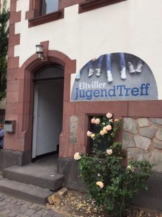Eingang zum JUZ Eltville. Eingang Jugendzentrum: Vor dem Eingang blühen Rosen und es hängt ein Schild "Eltviller Jugendzentrum" am Gebäude.