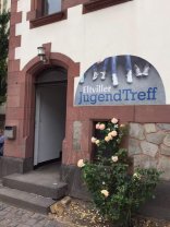 Eingang zum JUZ Eltville. Eingang Jugendzentrum: Vor dem Eingang blühen Rosen und es hängt ein Schild "Eltviller Jugendzentrum" am Gebäude.