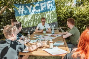Das Team von YCFF trifft sich regelmäßig zum Austausch. Jugendliche sitzen an einem Gartentisch im Schatten und diskutieren. Im Hintergrund hängt eine Flagge der Gemeinschaft "Your City For Future".