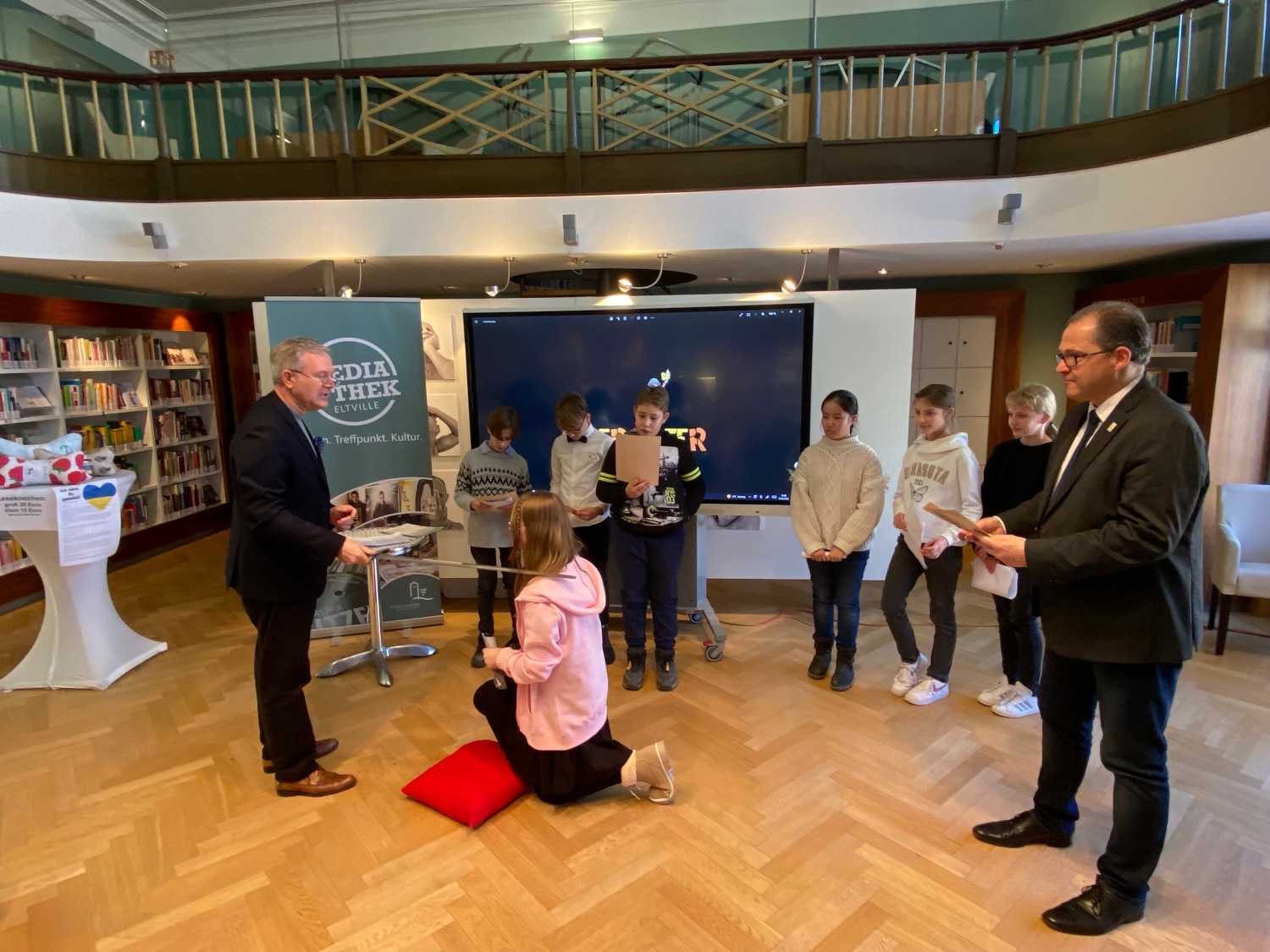 Ein Mädchen kniet auf einem Kissen, während der Schulleiter sie mit einem Schwert zum Leseritter schlägt. Die anderen Kinder und Bürgermeister Kunkel stehen nebendran