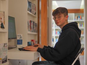 In der Mediathek gibt es zwei Internetplätze, die genutzt werden dürfen. Ein Jugendlicher schaut in die Kamera und sitzt vor einem Computer.