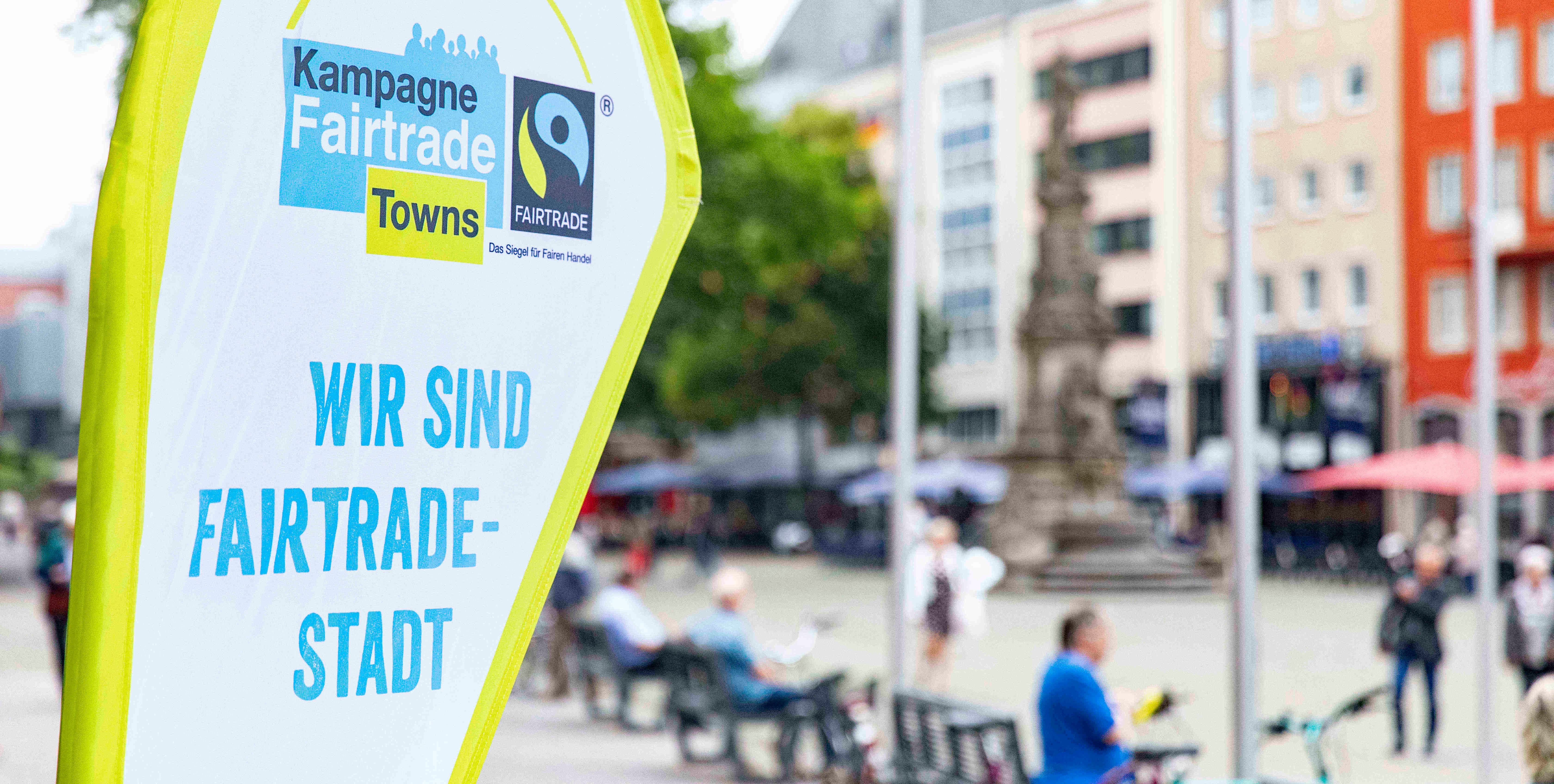 Fairtrade-Banner links, im Hintergrund befinden sich verschwommen Fußgänger, Radfahrer und ein Turm.