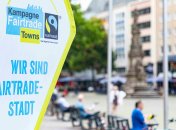 Fairtrade-Banner links, im Hintergrund befinden sich verschwommen Fußgänger, Radfahrer und ein Turm.