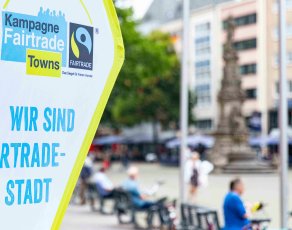 Fairtrade-Banner links, im Hintergrund befinden sich verschwommen Fußgänger, Radfahrer und ein Turm.