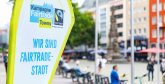 Eltville ist Fairtrade Town. Fairtrade-Banner links, im Hintergrund befinden sich verschwommen Fußgänger, Radfahrer und ein Turm.
