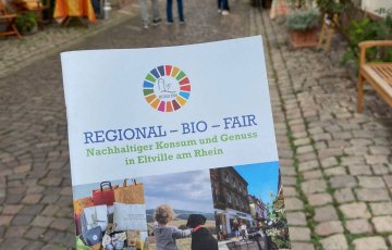 Jemand hält den nachhaltigen Einkaufsratgeber in der Hand, im HIntergrund sieht man die Eltviller Altstadt.