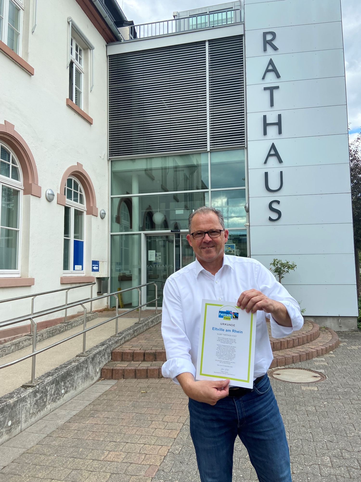 Bürgermeister Kunkel steht mit der Urkunde Fairtrade-Town vor dem Rathaus in Eltville.