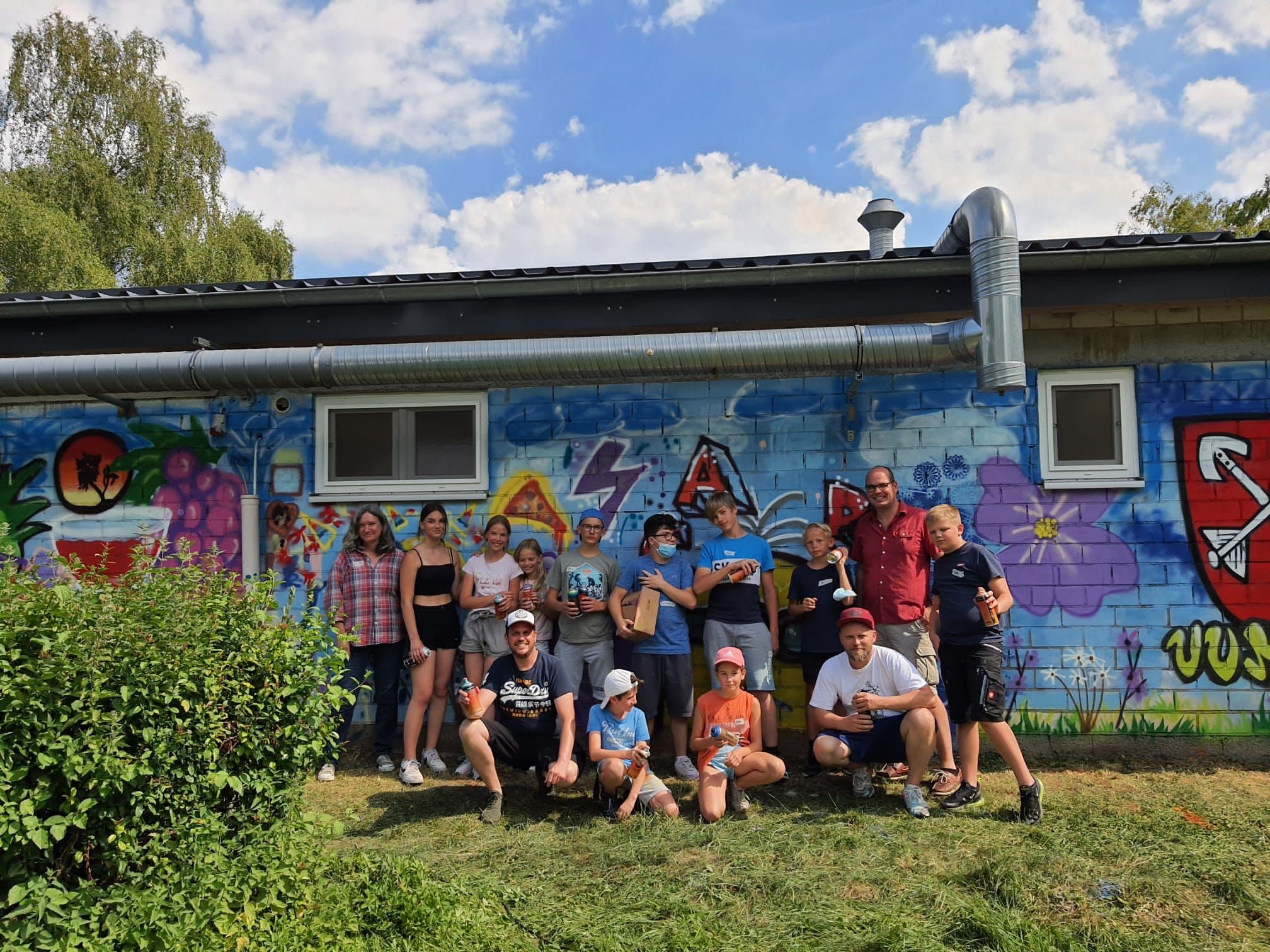 Gruppenfoto der teilnehmenden Kinder und Jugendlichen sowie Organisatoren vor der fertig besprühten Wand.