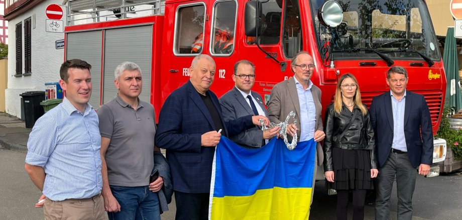 Vertreter der Helping Hands, Bügermeister Kunkel und die Delegation aus der Ukraine stehen vor dem übergebenen Feuerwehrtanker und halten eine Ukraine-Flagge in der Hand.