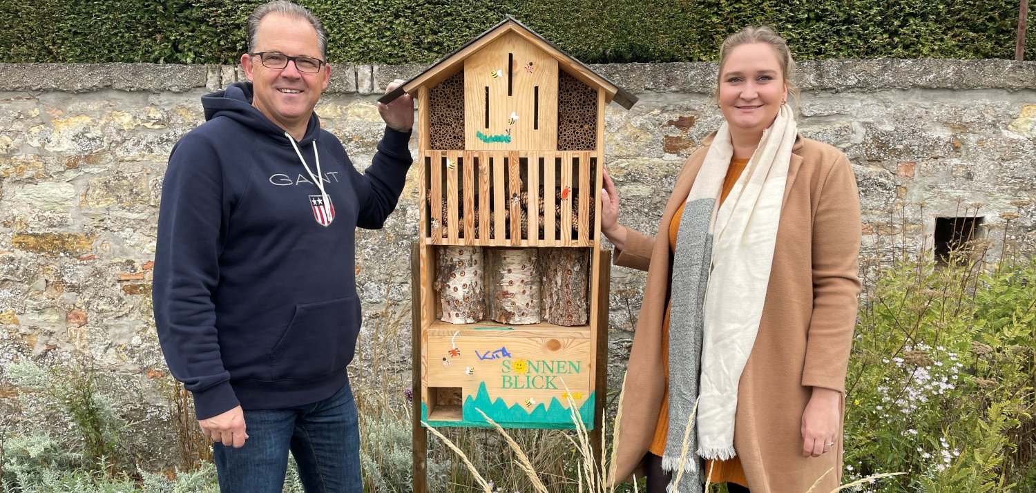 Bürgermeister Kunkel und Kitaleitung Sarah Hoffmann stehen neben dem Insektenhotel im Blumenbeet am Rheinufer.