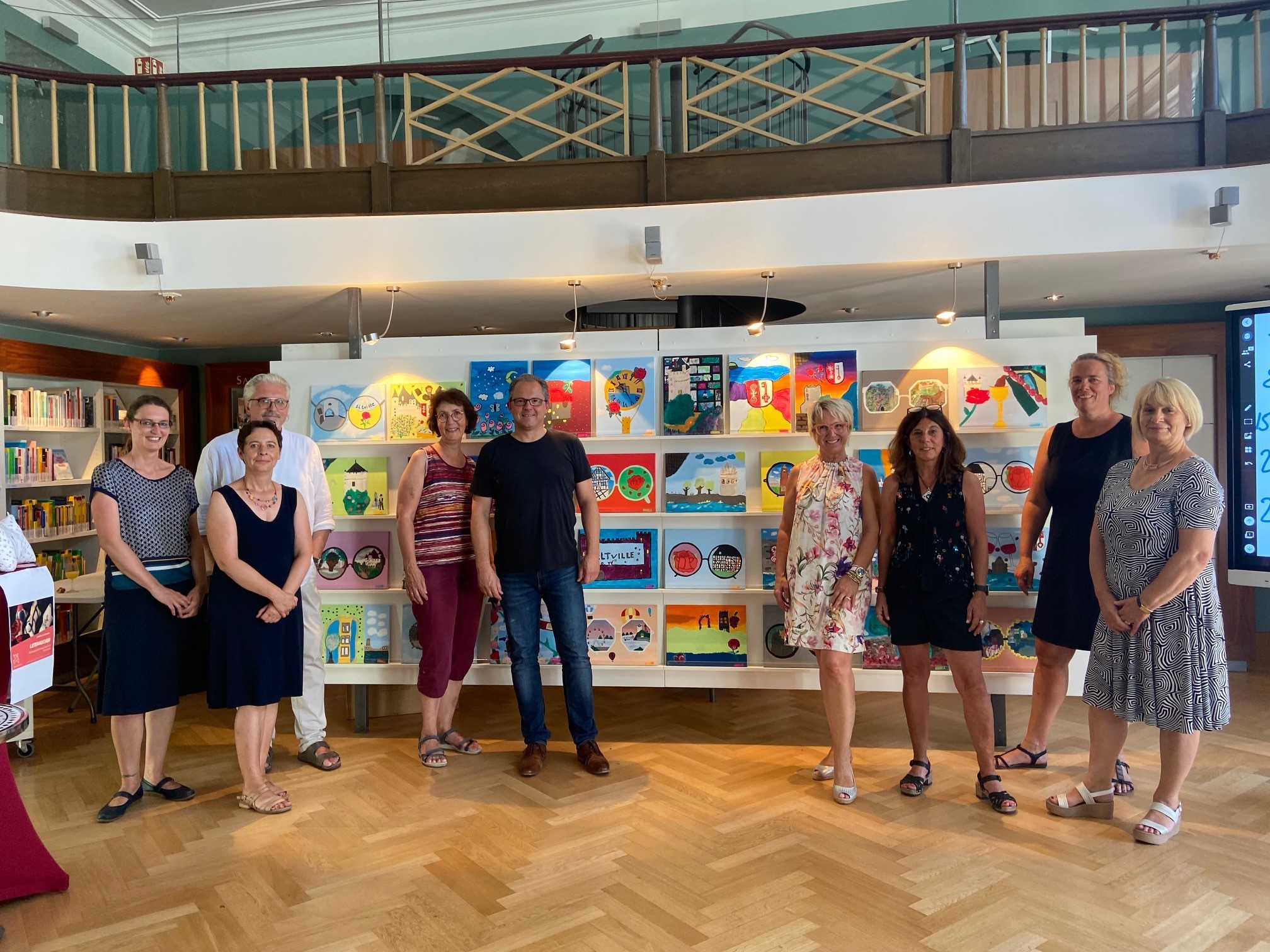 Die Jury aus Bürgermeister und Mitarbeitenden der Stadtverwaltung, Vertretern des Handels, des Fairtrade Steuerungskreises und der Realschule vor den gemalten Motiven der Realschüler, die in der Mediathek aufgestellt wurden. 