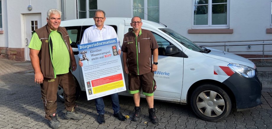 Bürgermeister Patrick Kunkel hält ein Plakat zur roten Wasserampel in der Hand, Ralph Biel (links) und Matthias Russ (rechts) von den Stadtwerken stehen neben ihm.