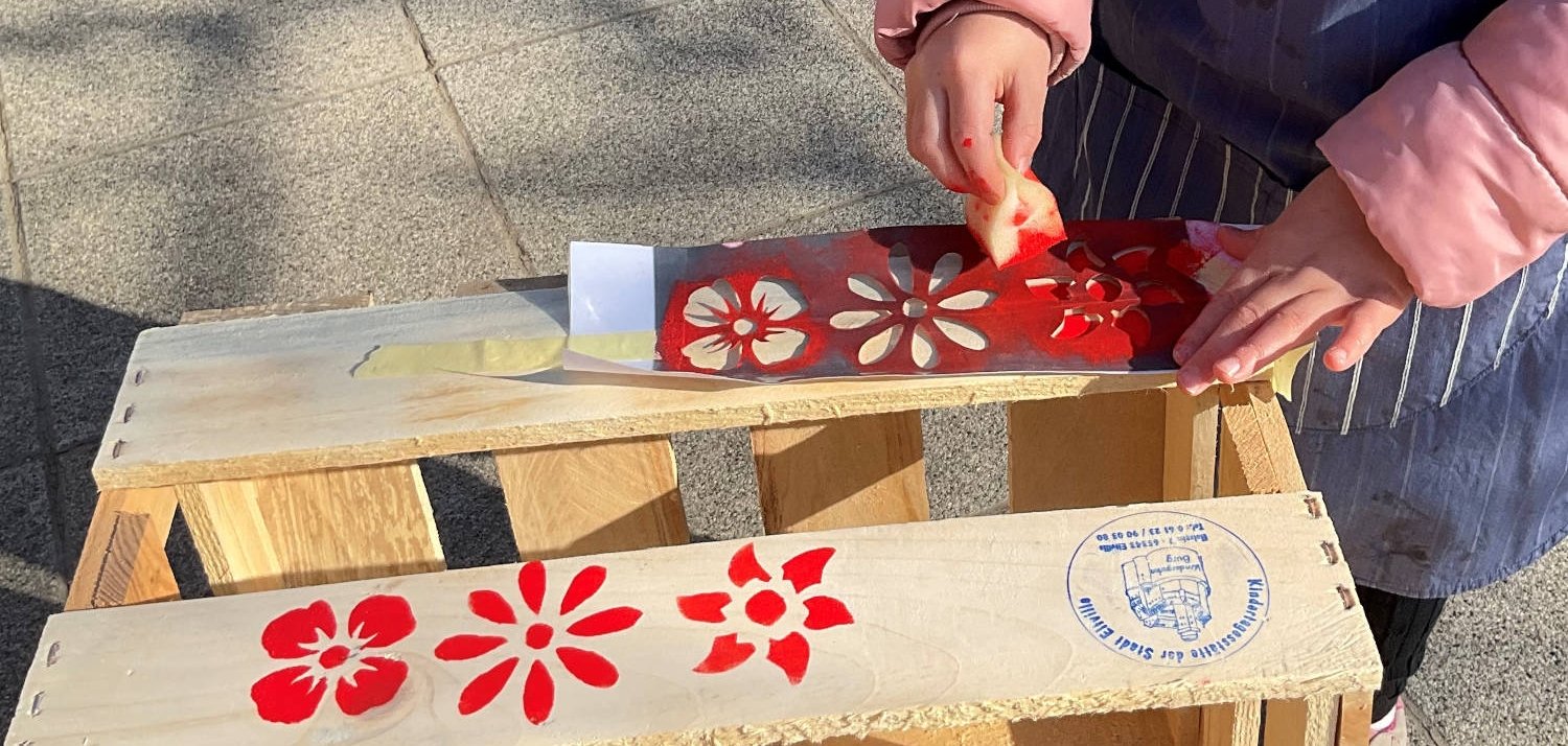Nach dem Abschleifen haben die Kids „ihre“ Kiste liebevoll gestaltet. Kinderhände stempeln rote Blumen auf eine kleine Holzkiste.