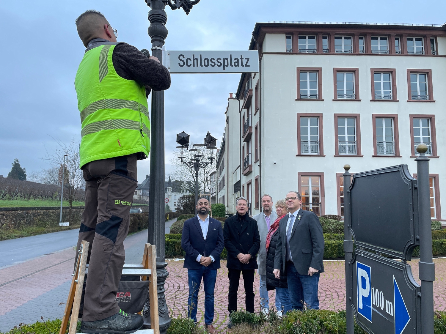Montage des neuen Straßenschildes durch einen Mitarbeiter der Stadtwerke unter Begutachtung des Bürgermeisters, der Ortsvorsteherin und Herren vom Schloss Reinhartshausen