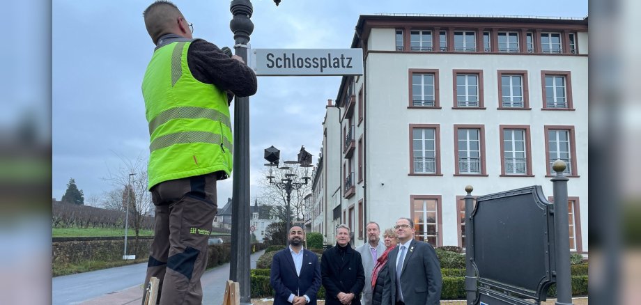 Montage des neuen Straßenschildes durch einen Mitarbeiter der Stadtwerke unter Begutachtung des Bürgermeisters, der Ortsvorsteherin und Herren vom Schloss Reinhartshausen