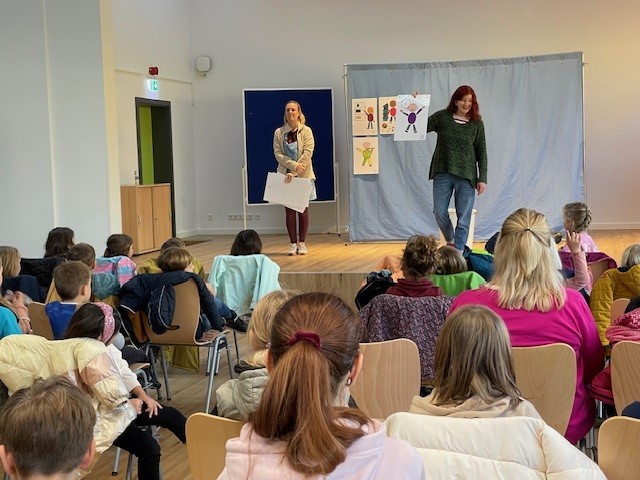 Zwei Frauen stehen auf einer Bühne, mehrere Kinder schauen zu, man sieht diese von hinten