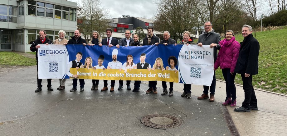 Mitglieder des Runden Tisches "Arbeitskräftemangel Rheingau" sowie Vertreter der Rheingauer Kommunen und des Dehoga bringen ein Banner zur Kampagne "Arbeitskräftegewinnung" zum Eltviller Schulzentrum.