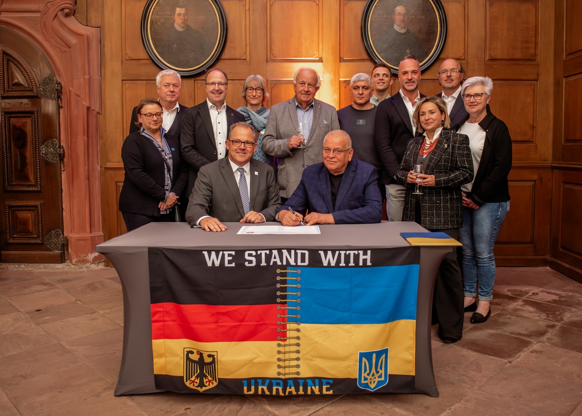 An einem Tisch sitzen die Bürgermeister aus Eltville und Zwenyhorodka. Hinter ihnen stehen 4 Frauen und 7 Männer aus den jeweiligen Delegationen der beiden Städte. Vorne am Tisch hängt eine kombinierte Deutsch-Ukrainische Flagge. Auf dieser steht: We stand with Ukraine.