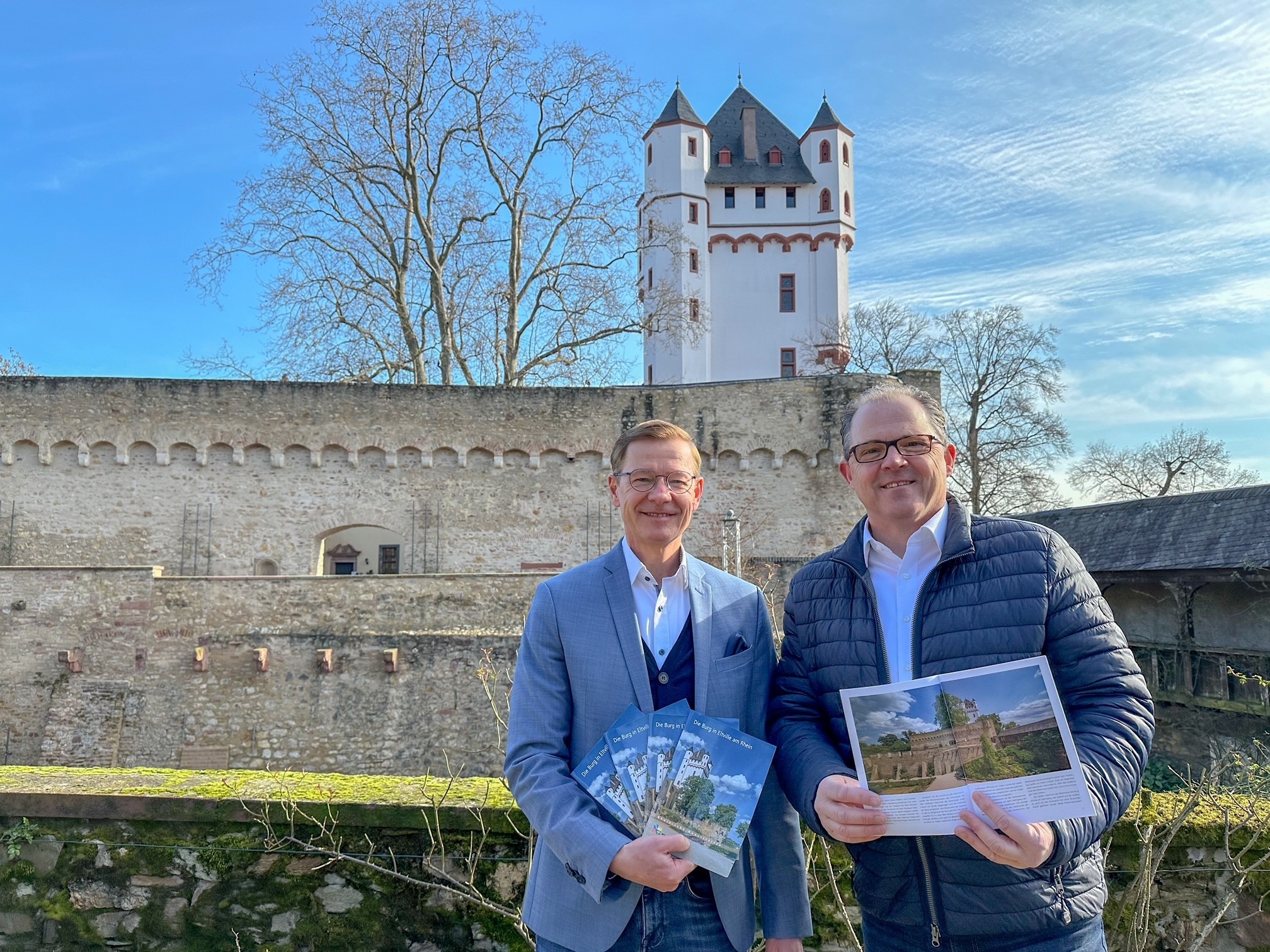Bürgermeister Patrick Kunkel und Geschäftsführer des Burgvereins, Thomas März, präsentieren die neue Borschüre über das Eltviller Wahrzeichen vor der Kurfürstlichen Burg selbst