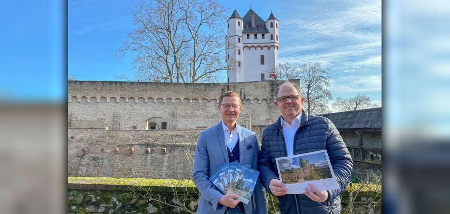 Bürgermeister Patrick Kunkel und Geschäftsführer des Burgvereins, Thomas März, präsentieren die neue Borschüre über das Eltviller Wahrzeichen vor der Kurfürstlichen Burg selbst