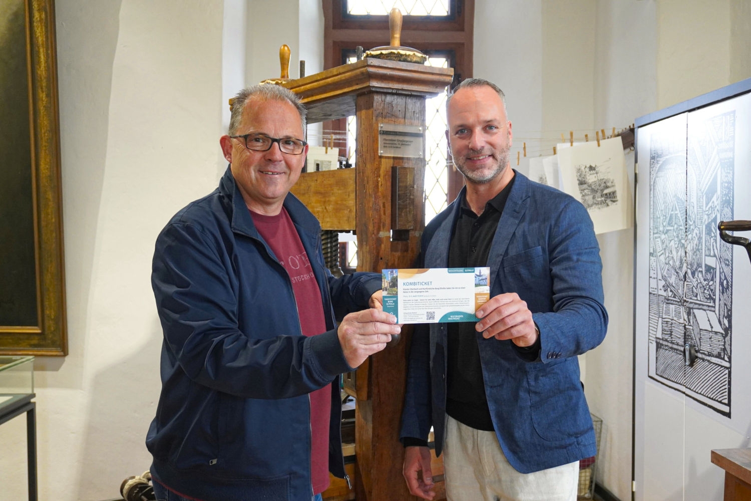 Bürgermeister Patrick Kunkel (links) und Julius Wagner, Vorstandsvorsitzender der Stiftung Kloster Eberbach, präsentieren das neue Kombiticket für den Besuch von Kloster Eberbach und Kurfürstlicher Burg. Die beiden stehen vor der historischen Druckerpresse – einem Kleinod in der Eltviller Burg.