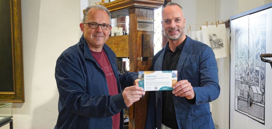 Bürgermeister Patrick Kunkel (links) und Julius Wagner, Vorstandsvorsitzender der Stiftung Kloster Eberbach, präsentieren das neue Kombiticket für den Besuch von Kloster Eberbach und Kurfürstlicher Burg. Die beiden stehen vor der historischen Druckerpresse – einem Kleinod in der Eltviller Burg.